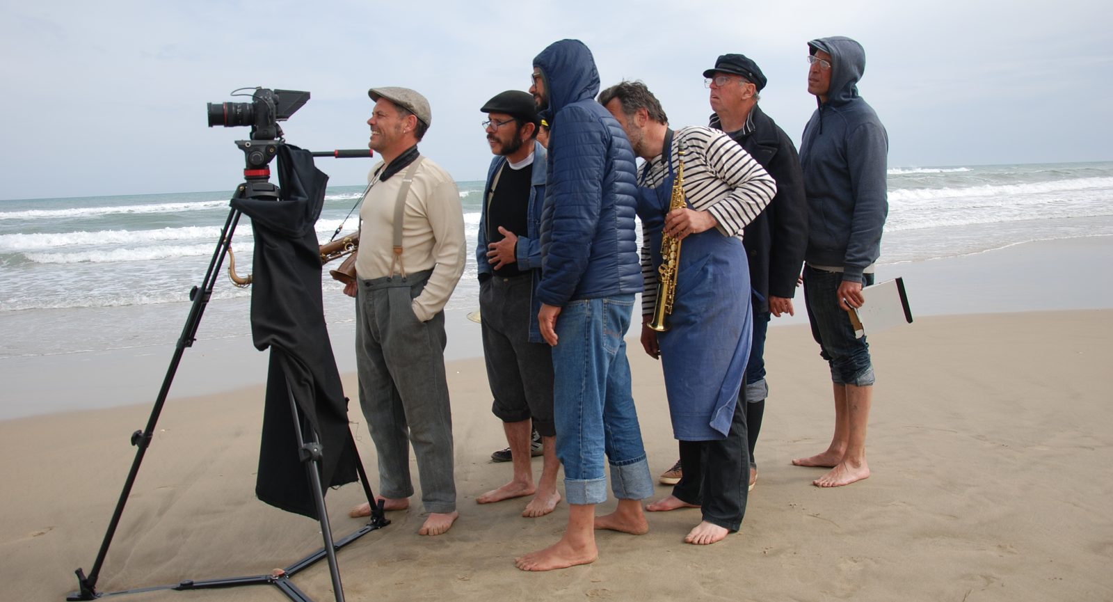 Barefoot on a beach, crew members and musicians, saxophones in hand, crowd around the camera facing the sea